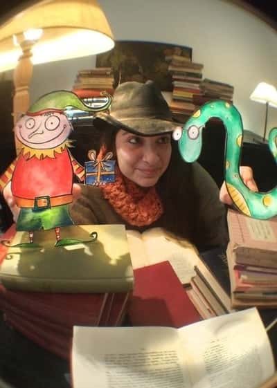 Η Αγγελική φοράει καπέλο και διαβάζει βιβλίο μαζί με ζωγραφισμένους χαρακτήρες.